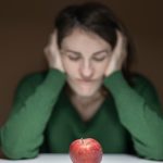 A woman in a green sweater observes an apple on a table, creating a thoughtful scene.