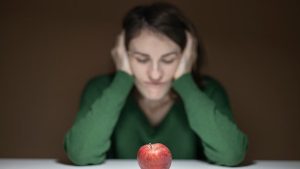 A woman in a green sweater observes an apple on a table, creating a thoughtful scene.