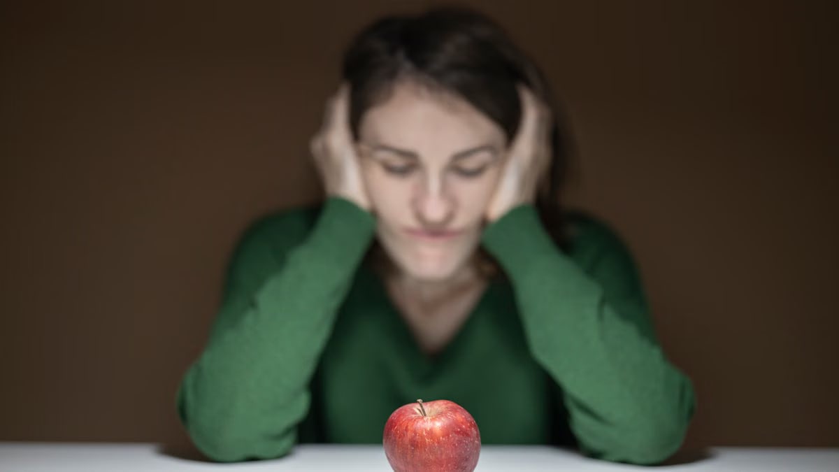 A woman in a green sweater observes an apple on a table, creating a thoughtful scene.